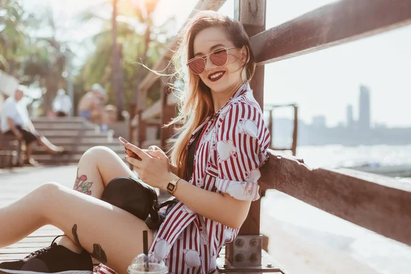 Усміхнена молода жінка використовує смартфон, сидячи на пірсі на пляжі і дивлячись на камеру — стокове фото