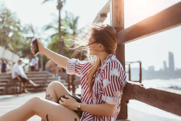 Beautiful young woman takign selfie while sitting on floor of pier at beach — Stock Photo