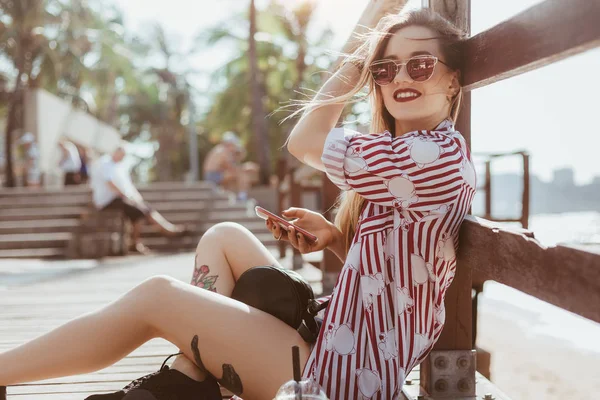 Seitenansicht der schönen jungen Frau, die auf dem Fußboden eines hölzernen Pier sitzt und das Smartphone benutzt — Stockfoto