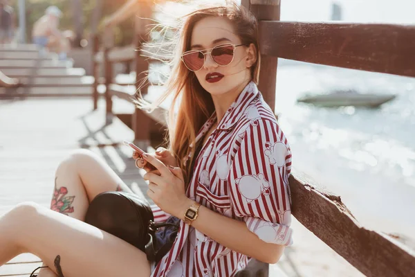 Side view of young woman sitting on floor of wooden pier and using smartphone — Stock Photo