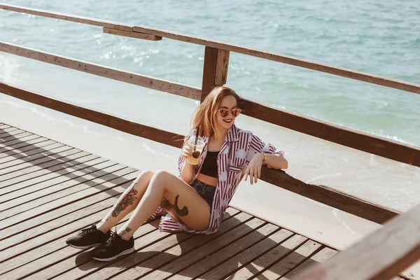 Bela jovem mulher com copo de plástico sentado no cais de madeira na frente do mar — Fotografia de Stock