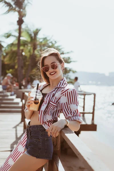 Mujer joven sonriente con taza de plástico de cóctel apoyado de nuevo en el muelle en la playa - foto de stock