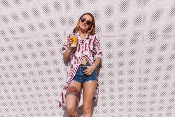 Attractive young woman with mango shake in plastic cup leaning back on wall — Stock Photo