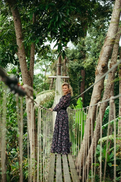 Sorridente donna attraente in piedi sul ponte pedonale nella giungla e guardando altrove — Foto stock