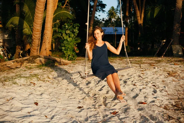 Усміхнена красива жінка на гойдалках між пальмами дивиться на камеру на пляжі — стокове фото
