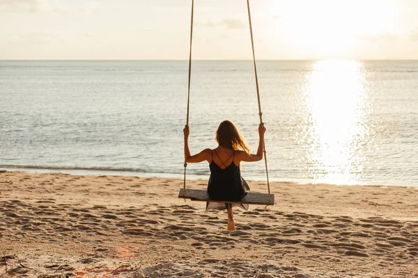 Visão traseira da mulher no balanço na praia do oceano durante o pôr do sol — Fotografia de Stock