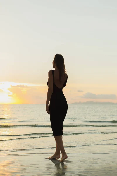 Вид ззаду приваблива жінка стоїть босоніж на піщаному пляжі океану під час заходу сонця — стокове фото