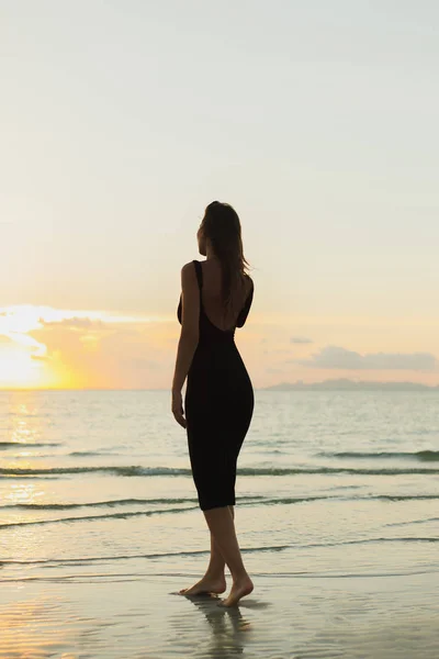 Vista trasera de la atractiva mujer de pie descalza en la playa de arena del océano durante el atardecer - foto de stock