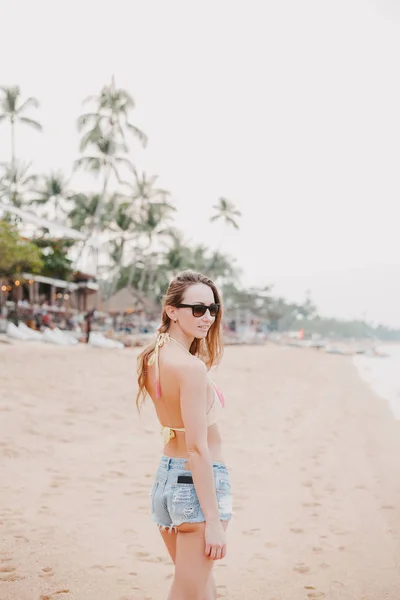 Attractive girl standing on sandy beach in bikini top and sunglasses — Stock Photo
