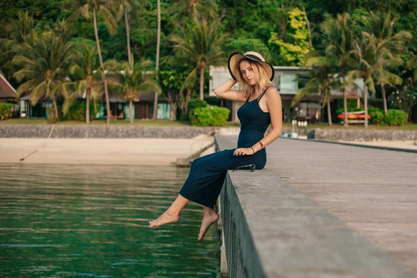 Seitenansicht der schönen Frau mit Hut und Kleid, die auf der Seebrücke sitzt und in die Kamera schaut — Stockfoto