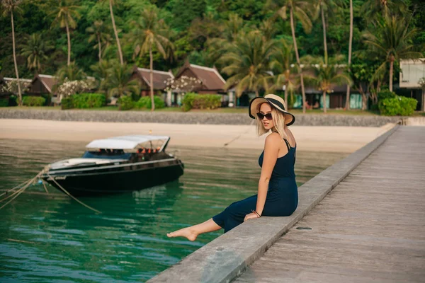 Vista lateral de la hermosa mujer en sombrero y vestido sentado en el muelle cerca del océano - foto de stock