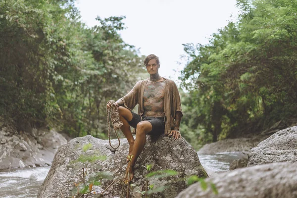 Tätowierter Mann mit Wasserflasche ruht auf Felsen mit grünen Pflanzen und Fluss im Hintergrund, bali, Indonesien — Stockfoto