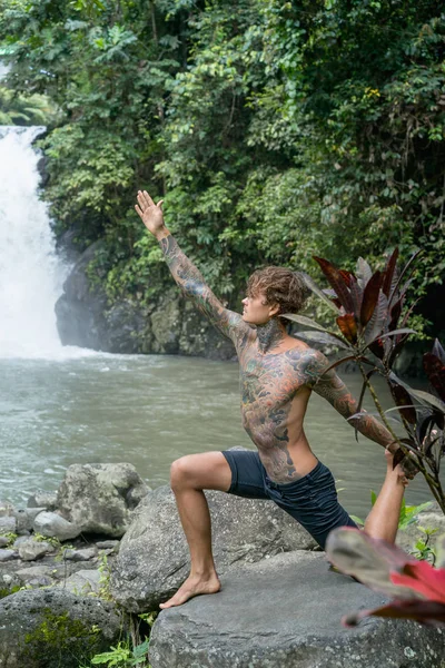 Tattooed man practicing yoga on rock with Aling-Aling waterfall and green plants on background, Bali, Indonesia — Stock Photo