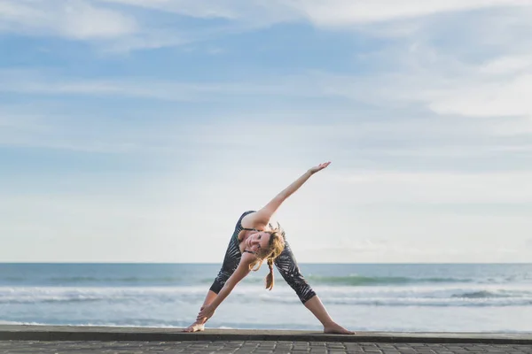 Femme dans la pose de yoga triangle étendu avec l'océan et le ciel bleu sur fond, Bali, Indonésie — Photo de stock