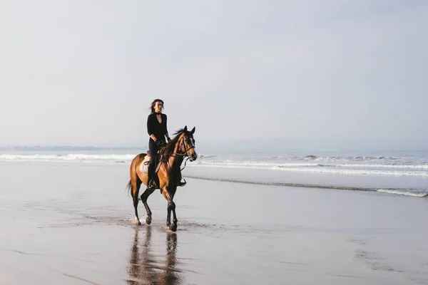 Mulher equitação cavalo na praia arenosa com oceano atrás — Fotografia de Stock