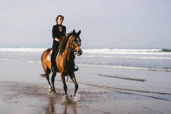 Jeune femme équitation cheval avec l'océan ondulé derrière — Photo de stock