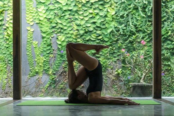 Side view of athletic young woman practicing yoga in entrance into Plow pose (Halasana) in front of wall covered with green leaves — Stock Photo