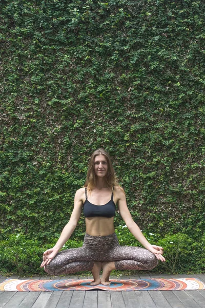 Fit young woman practicing yoga in Squatting Toe Balance pose making Gyan Mudra in front of wall covered with green leaves — Stock Photo