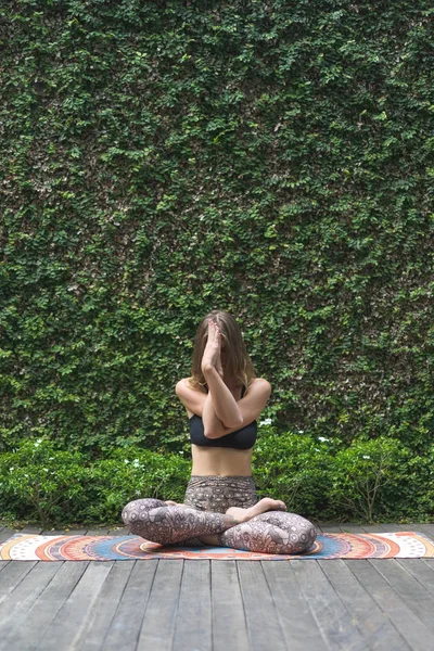 Hermosa joven practicando yoga en pose de loto frente a la pared cubierta de hojas verdes - foto de stock