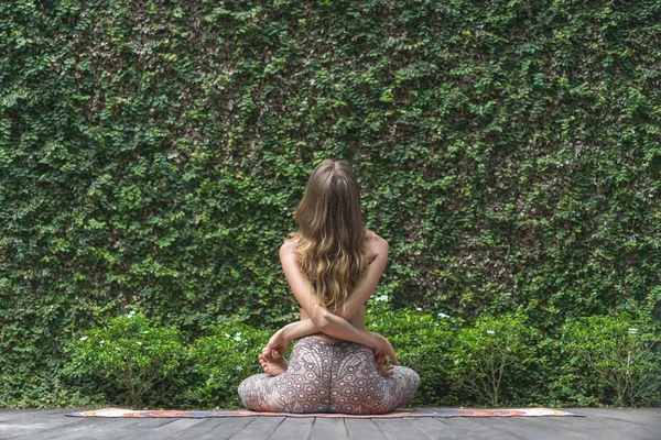 Rear view of woman practicing yoga in lotus pose in front of wall covered with green leaves — Stock Photo