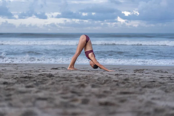 Seitenansicht einer attraktiven jungen Frau, die Yoga in abwärts gerichteter Hundepose (adho mukha svanasana) an der Küste praktiziert — Stockfoto