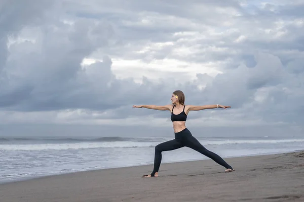 Vista lateral de una hermosa joven practicando yoga en pose de Guerrero (Virabhadrasana) en la orilla del mar - foto de stock