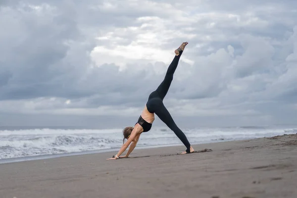 Joven deportista practicando yoga en pose One Legged Downward-Facing Dog (Eka Pada Adho Mukha Svanasana) a orillas del mar - foto de stock