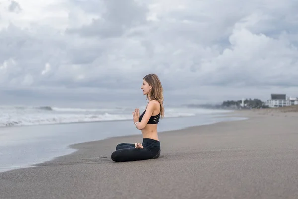 Vista laterale di bella giovane donna che pratica yoga in posa di loto (padmasana) con mudra namaste in riva al mare — Foto stock