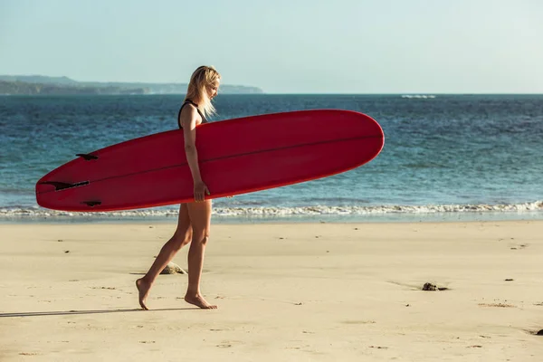 Surfista femenina sosteniendo tabla de surf y caminando en la playa cerca del mar - foto de stock