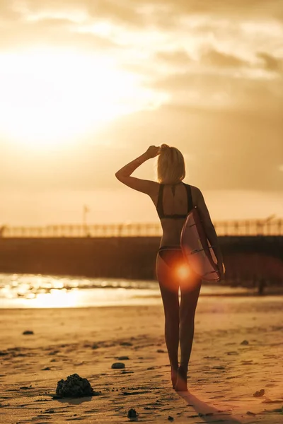 Vista trasera de la niña posando con tabla de surf en la playa al atardecer con luz de fondo - foto de stock