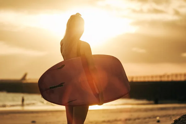 Visão traseira do surfista posando com prancha na praia ao pôr do sol com retroiluminação — Fotografia de Stock