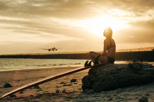 Mulher sentada na rocha com prancha de surf na praia ao pôr do sol com avião no céu — Fotografia de Stock