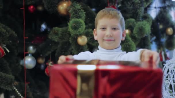 Happy Smileing Boy показывает красный новогодний подарок. Мальчик стоит под елкой — стоковое видео