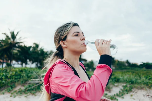 Fitness runner woman drinking water of a sport bottle. Athlete girl taking a break during run to hydrate during hot summer exercise on beach. Healthy active lifestyle. — Stock Photo, Image