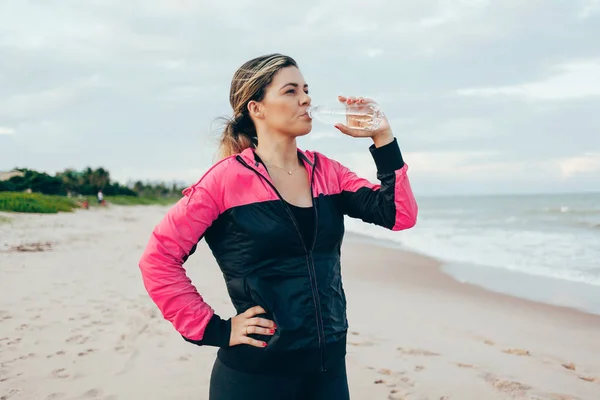 Corredor de fitness mujer bebiendo agua de una botella deportiva. Atleta chica tomando un descanso durante la carrera para hidratarse durante el calor del ejercicio de verano en la playa. Vida activa saludable . — Foto de Stock