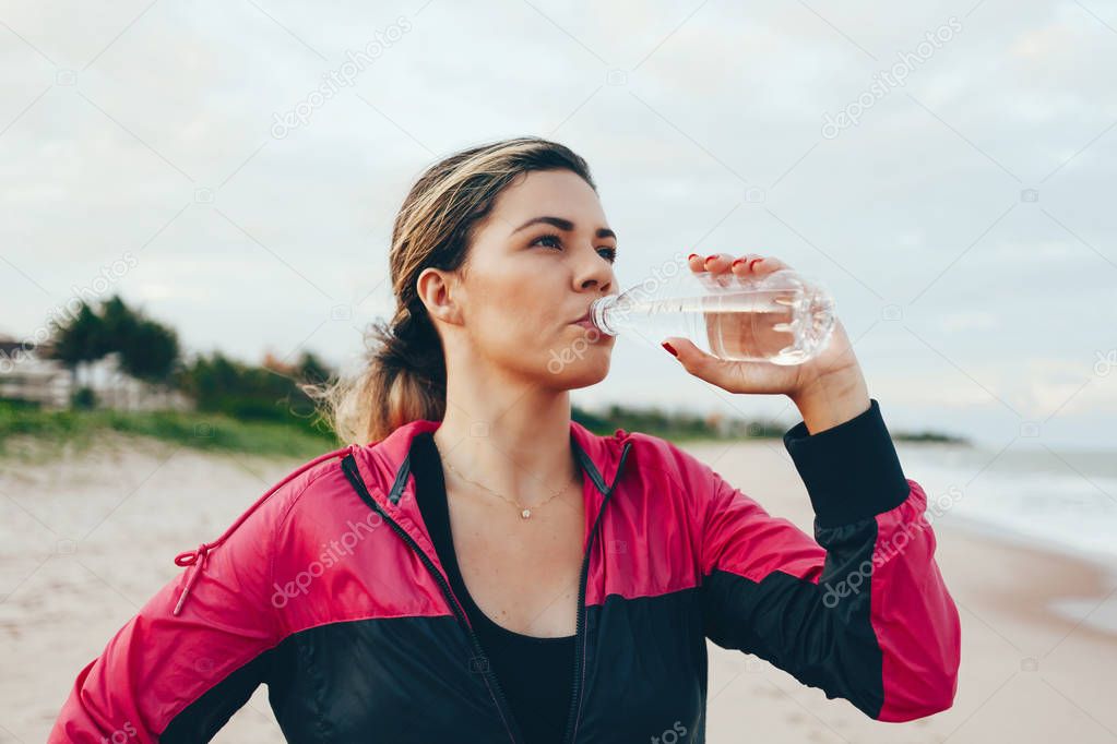 Fitness runner woman drinking water of a sport bottle. Athlete girl taking a break during run to hydrate during hot summer exercise on beach. Healthy active lifestyle.