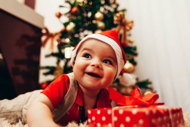 Bebek, arka planda Noel ağacı ışıkları eşliğinde oturma odasında hediye kutusuyla oynuyor.