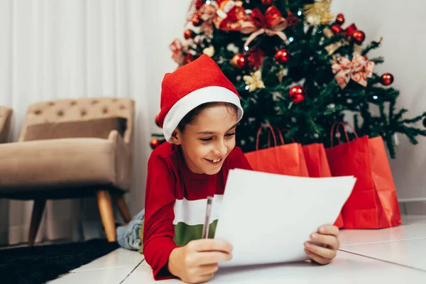 Anak itu tergeletak di lantai di bawah pohon Natal menulis daftar keinginan untuk Santa. Menunggu Natal. Perayaan. Tahun Baru . — Stok Foto