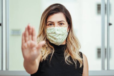 Yüzünde koruyucu maske takan Brezilyalı genç bir kadının elinde dur işareti var. Coronavirus COVID-19 önleme.