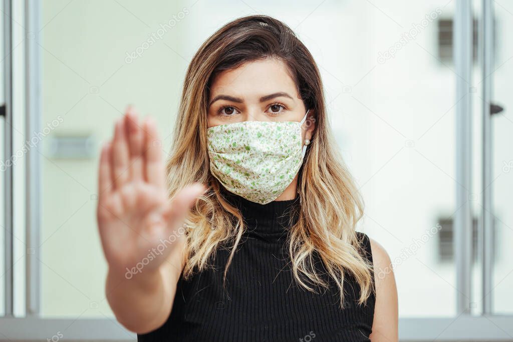 Brazilian young woman in protective mask on face showing stop sign with her hand. Coronavirus COVID-19 prevention.