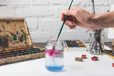 partial view of artist putting paint brush into glass jar with water at workplace clipart