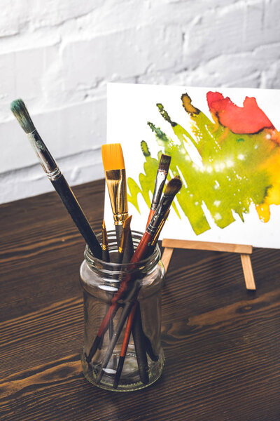 close-up view of various paint brushes in glass and abstract painting behind at designer workplace