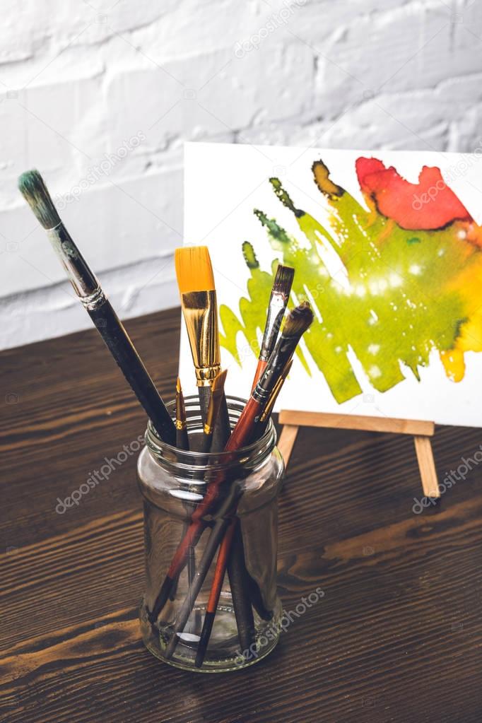 Close-up view of various paint brushes in glass and abstract painting behind at designer workplace