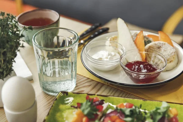 Close-up view of boiled egg, glass of water and cheesecakes with jam and sour cream for breakfast — Stock Photo