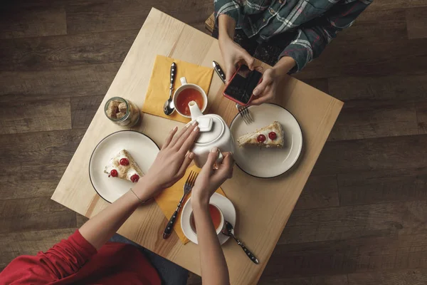 Top view of person pouring tea from kettle while friend using smartphone during breakfast — Stock Photo