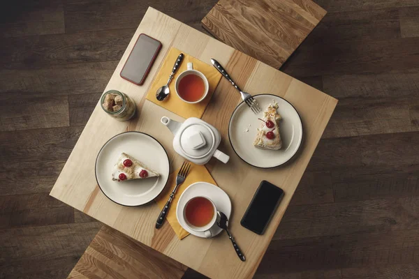 Vista superior de la tetera, tazas de té, pasteles y teléfonos inteligentes con pantallas en blanco en la mesa - foto de stock