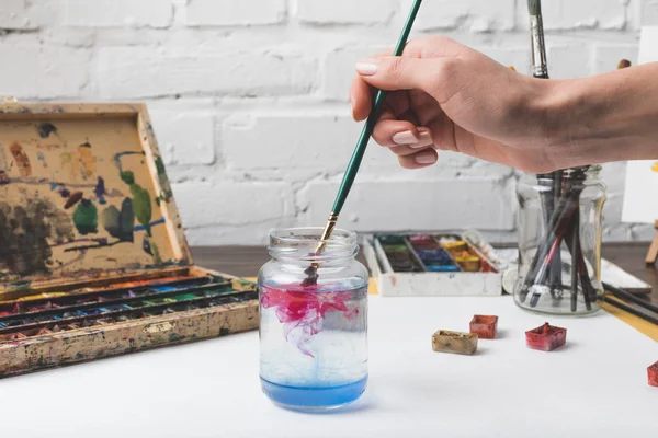 Vista parcial del artista poniendo pincel en frasco de vidrio con agua en el lugar de trabajo - foto de stock