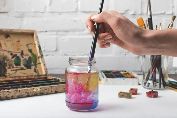 Recortado tiro de artista poniendo pincel en frasco de vidrio con agua en el lugar de trabajo - foto de stock