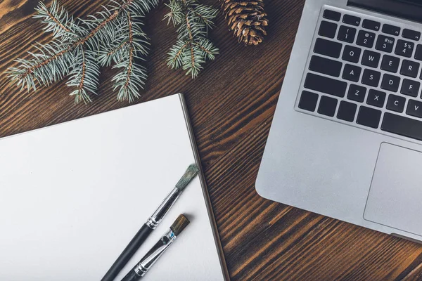 Vue rapprochée de l'ordinateur portable, album de dessin vierge et pinceaux avec branches de sapin et cône de pin sur le lieu de travail du concepteur — Photo de stock