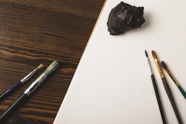 Vista superior de pincéis de pintura, concha e álbum de desenho em branco na mesa de madeira — Fotografia de Stock